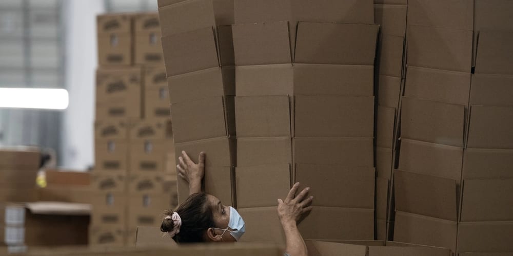 A worker carrying a tall stack of flat-packed cartons past staged boxes in a packing hall.