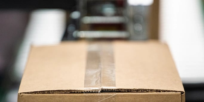 A sealed and taped carton on a packing line, with the line running out of focus behind it.