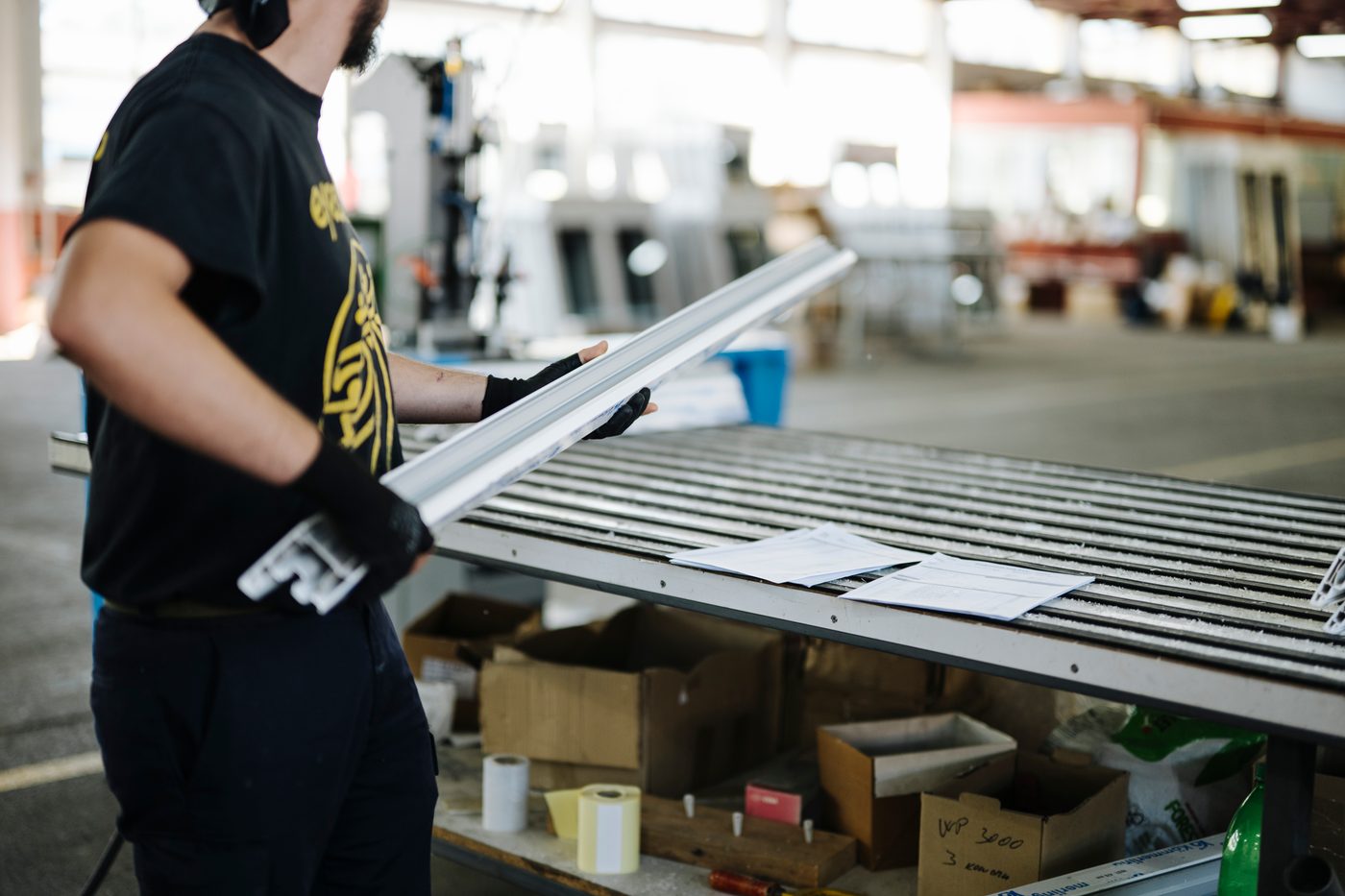 A worker in gloves handling a component at a staging table, with parts boxed in bins and build sheets laid out below.