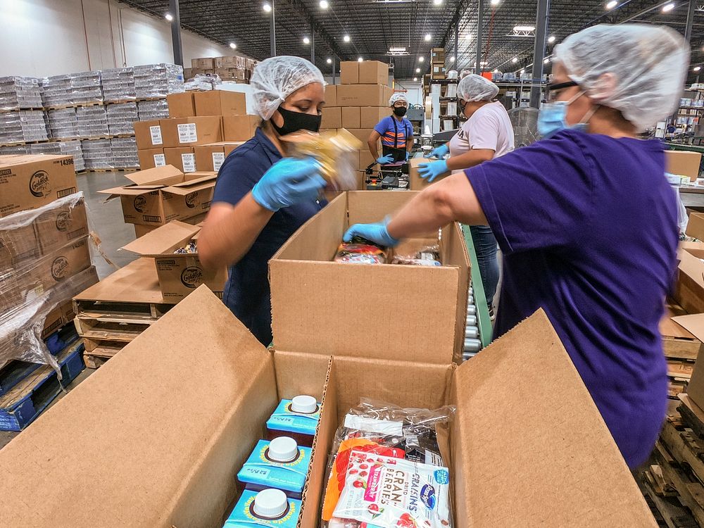 A packing crew filling cartons with product beside a conveyor in a distribution facility.
