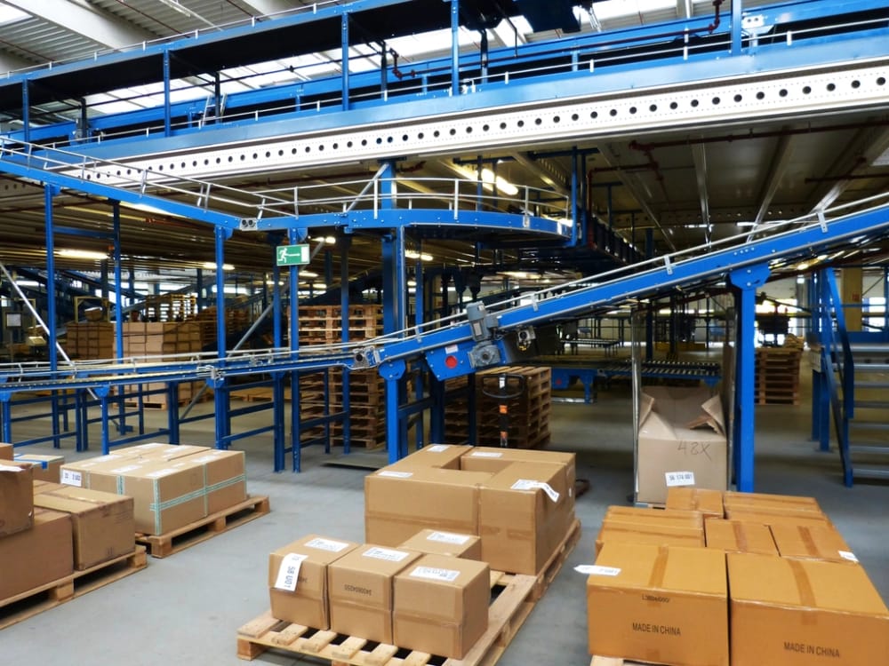 Returned cartons staged on pallets beside a conveyor in a receiving area, one box opened for triage.