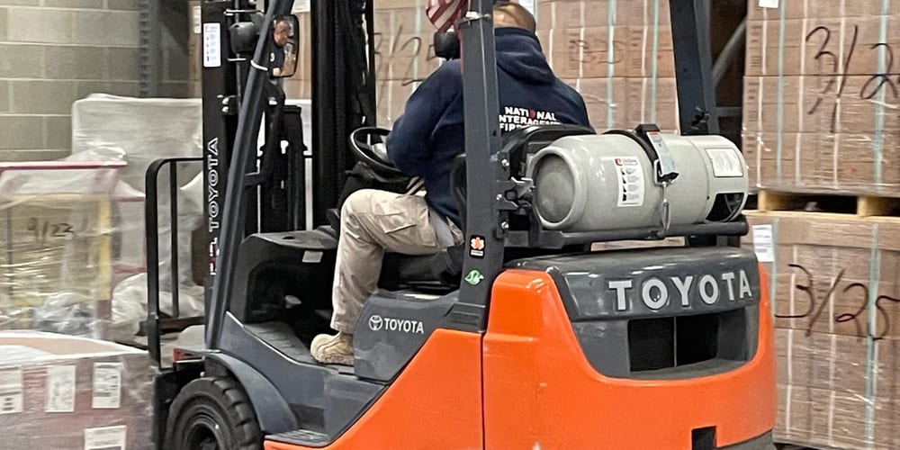 A forklift operator moving through a warehouse past wrapped pallets marked up with handwritten receipt dates.